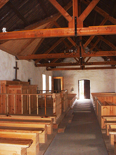 The simple interior of Tullyallen Masshouse shows the wooden altar in the middle of the back wall, open rafters and whitewashed walls. The confessional stands near the door. The building had a T-shape plan, the stem of the T being the sacristy and priest's house.